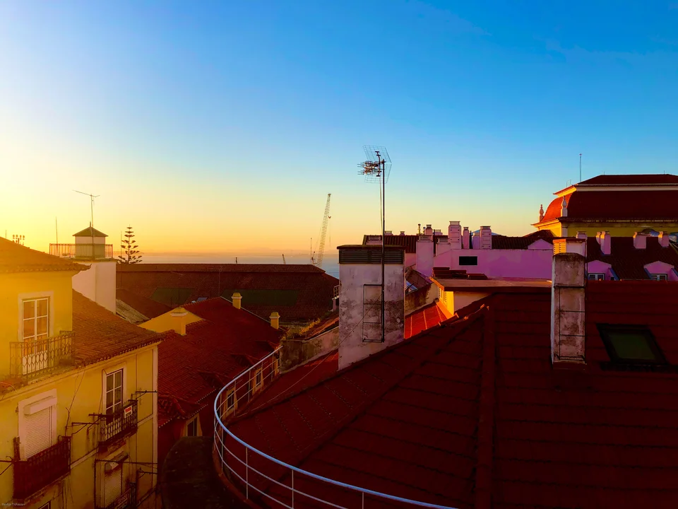 Lisbon rooftops and a curved metal railing in warm evening light beneath a clear blue sky.